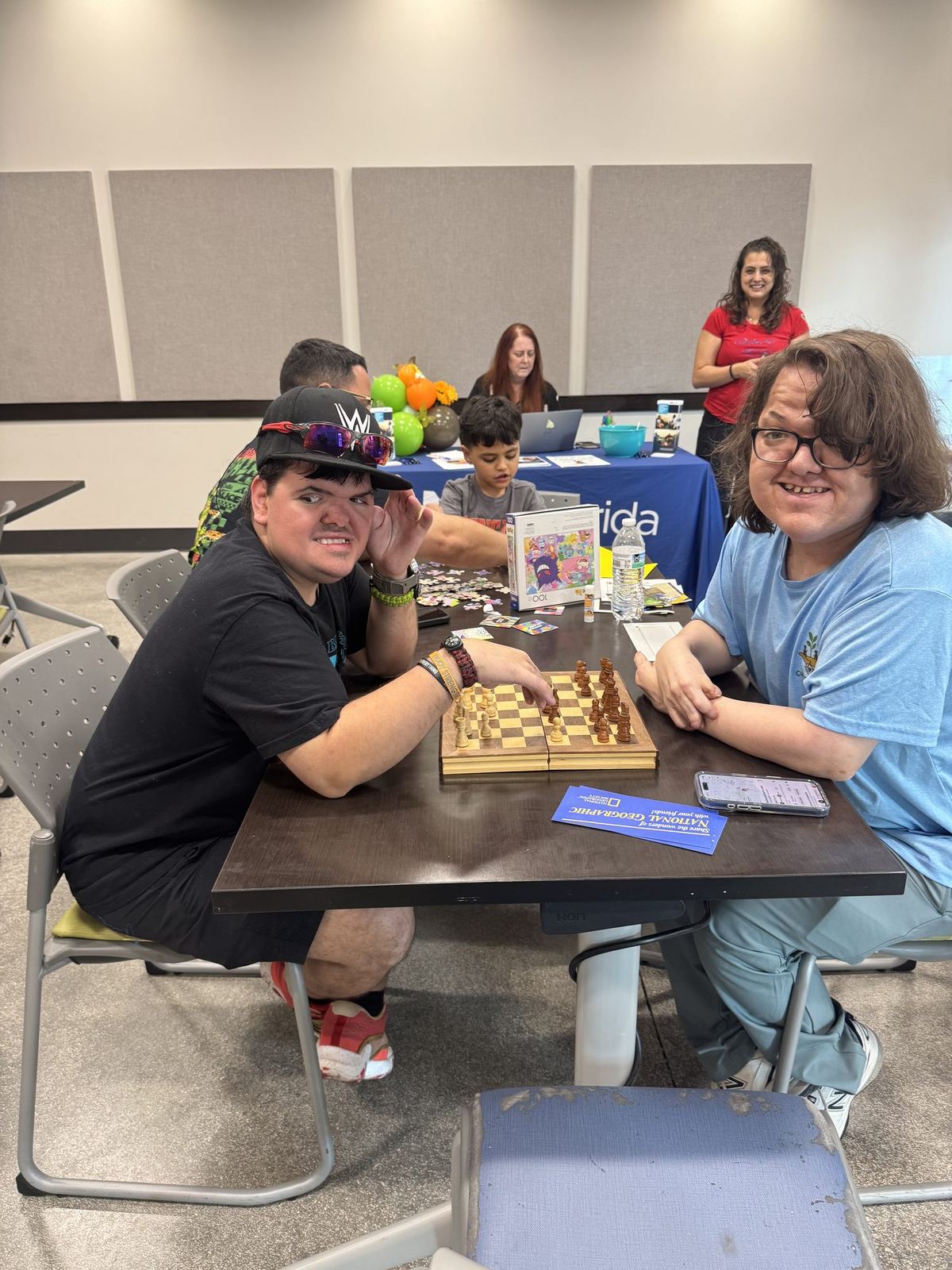 Two young ACES participants playing chess together at a community table while staff support others nearby
